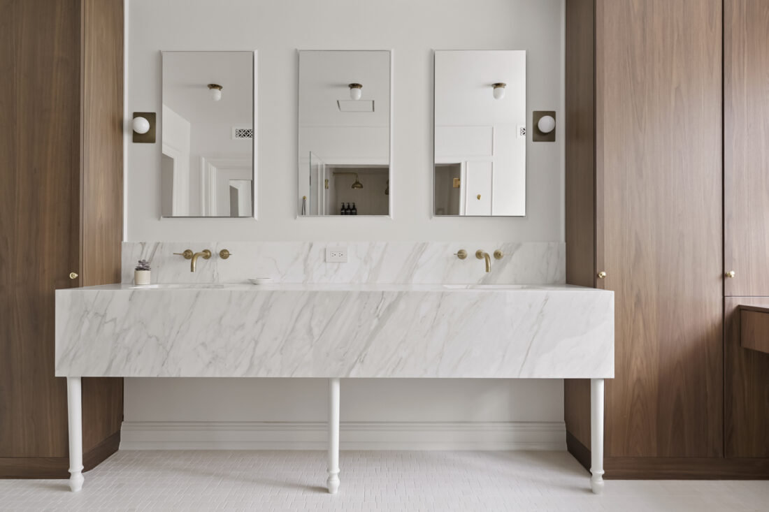 Marble double vanity on turned legs, framed by full-height walnut cabinetry, three mirrors and brass wall faucets.